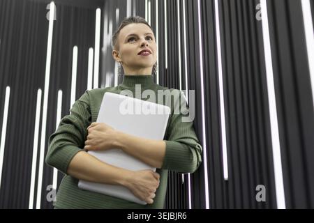 A mature woman programmer with a short haircut stands with a laptop in her hands against a black wall with backlights Stock Photo