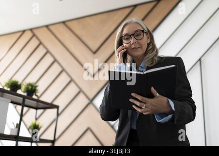 Mature businesswoman in glasses and business clothes speaks on a mobile phone with a notepad in her hands in the office Stock Photo