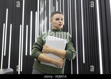 Successful woman programmer with a short haircut with a laptop in her hands against a black wall in the office Stock Photo