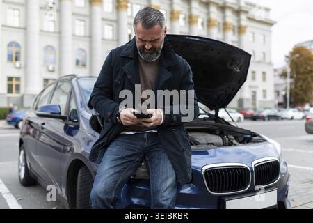 Adult man calls technical support for car repair next to a broken car with an open hood Stock Photo