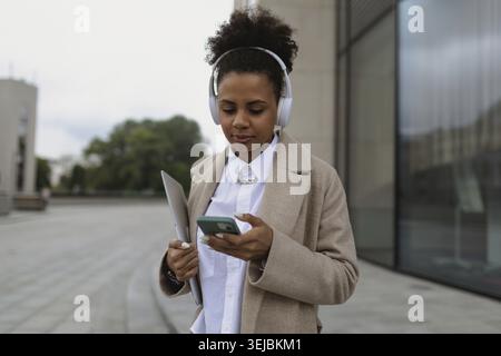 A young black female student in headphones with a mobile phone and a laptop walks around the city next to a modern building Stock Photo
