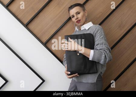 A business woman with a short haircut in a gray sweater with a leather folder for documents in her hands on the background of an office texture wall Stock Photo
