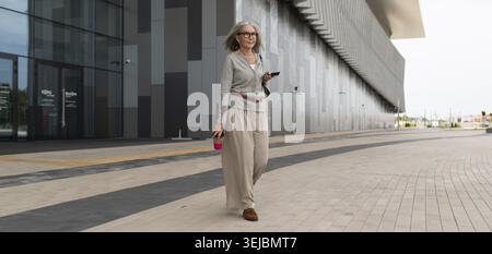 A mature woman strolls on a stylish plaza surrounded by contemporary architecture. She holds a coffee cup and appears relaxed while checking her phone Stock Photo