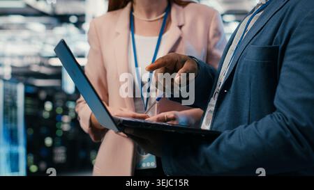 Engineering team solving tasks in server farm using laptop, doing network management virtualization. Teamworking data center colleagues oversee IT infrastructure supercomputers with notebook, camera B Stock Photo