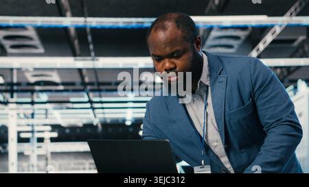 Programmer at server farm workplace desk using laptop, doing network management. Data center IT expert overseeing IT infrastructure supercomputers with software, typing on notebook keyboard, camera B Stock Photo