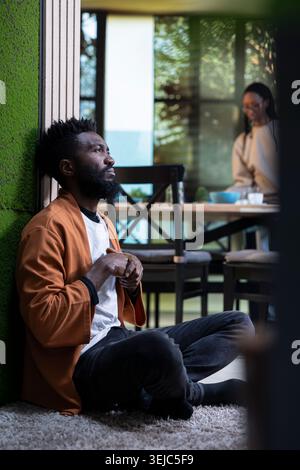 Black man, depressed and unwell, sitting alone, on the floor at home, surrounded by darkness. Person expressing sadness and vulnerability, mental health suffering linked to disappointment. Stock Photo