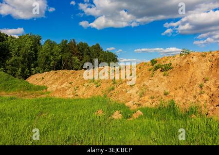 Large heaps of excavated sand and raw dirt piles disrupt a lush green field near a dense forest, highlighting the impact of human industrial activity Stock Photo