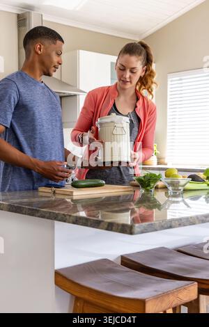 Couple cooking at kitchen island, man in blue tee, woman in coral jacket inspecting compost pail Stock Photo