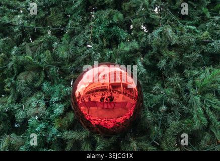Large red Christmas ornament hangs on an artificial pine tree and reflects the Suria KLCC mall plaza and visitors in Kuala Lumpur, Malaysia Stock Photo