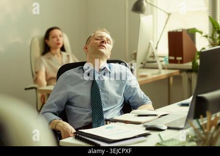 Middle aged Caucasian businessman leaning back with closed eyes at desk, feeling exhausted during office work. Suitable for burnout, workplace stress, fatigue awareness campaigns Stock Photo