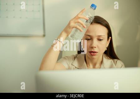 Young adult Caucasian businesswoman cooling forehead with water bottle while working on laptop, showing heat stress and fatigue. Useful for workplace wellness, summer office safety Stock Photo