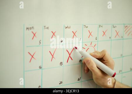Close up of adult hand marking wall calendar with red pen, crossing off days and planning schedule. Suitable for time management, deadline tracking, productivity campaign use Stock Photo