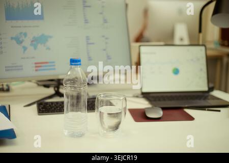 Close up of office desk showing water bottle and glass beside computer monitors displaying analytics, supporting workplace hydration, productivity and employee wellness content Stock Photo