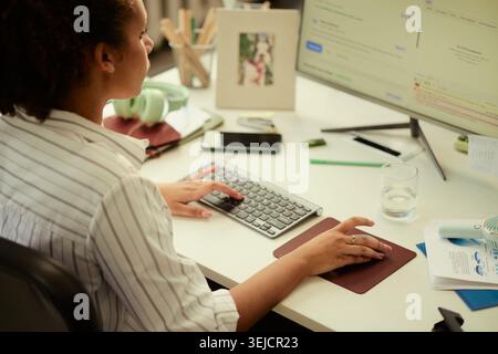 Young adult woman working at desktop computer, typing and navigating digital interface at desk, supporting remote work, online research, content creation or office productivity Stock Photo