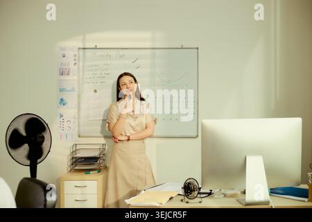 Young adult Asian businesswoman planning monthly strategy at whiteboard, holding marker and reviewing charts. Useful for business planning, project management, office workflow Stock Photo