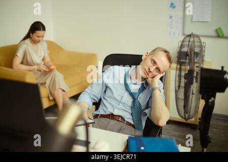 Young adult Caucasian businessman sitting exhausted at desk, leaning on hand near fan while coworker using smartphone in background. Office burnout concept for workplace stress campaigns Stock Photo