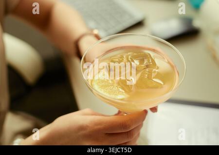 Close up of young adult woman holding iced citrus cocktail near laptop, suggesting after work break or social leisure. Suitable for hospitality marketing, lifestyle content, remote work balance Stock Photo