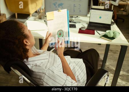 High angle view of young adult woman with disability cooling herself with handheld fan while reviewing documents at desk. Suitable for workplace accessibility, heat stress, remote work content Stock Photo