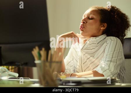 Young adult African American businesswoman cooling neck and exhaling while sitting at desk, showing workplace heat stress and fatigue. Useful for burnout, office wellness, summer work Stock Photo