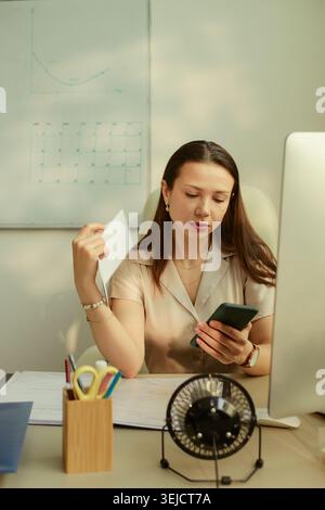 Young adult Caucasian businesswoman checking smartphone while holding document at desk, managing office workflow and communication. Useful for business productivity or admin content Stock Photo