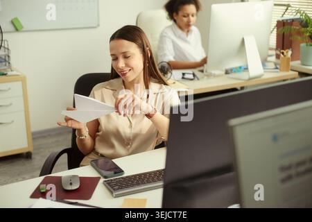 Young adult Caucasian businesswoman folding paper airplane at office desk, taking playful work break while colleague working behind. For teamwork, office culture, employee engagement marketing Stock Photo