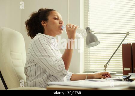 Young adult woman drinking water while working at desk, using computer during office break and supporting hydration, wellness, productivity. Side view for workplace health content Stock Photo
