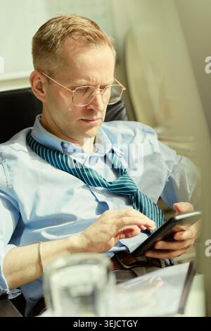 Vertical shot of Caucasian young adult businessman checking smartphone while sitting at workplace, managing mobile communication. Useful for business messaging or productivity content Stock Photo