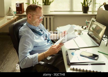 Caucasian middle aged businessman reviewing documents at desk beside laptop and smartphone, managing workload and office reporting. Useful for business productivity or deadline stress Stock Photo