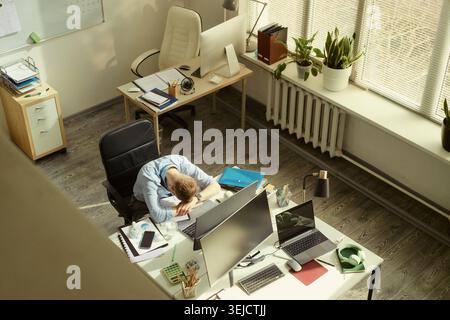 High angle shot of young adult Caucasian businessman resting head on desk beside computer monitors, showing workplace stress and burnout. Useful for mental health or overwork concepts Stock Photo