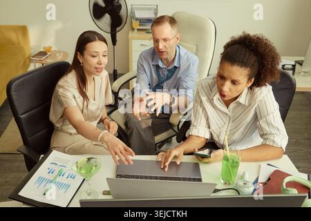 Diverse young adult coworkers reviewing laptop data, discussing project strategy, checking smartphone during office meeting. Useful for teamwork, startup planning, business communication Stock Photo
