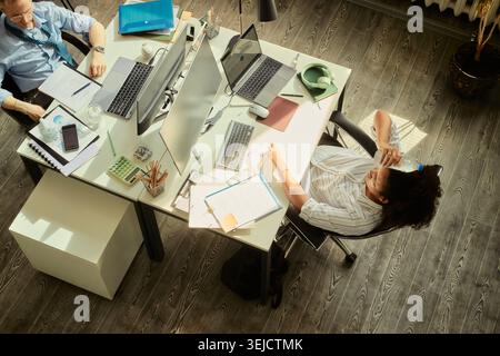 High angle view of Caucasian businessman and African American businesswoman working at shared desks, reviewing documents and laptops. Ideal for teamwork, office productivity, collaboration Stock Photo