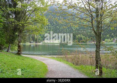 A scenic dirt path or gravel walkway leading towards a beautiful calm mountain lake surrounded by green trees. Peaceful summer day in an alpine Stock Photo