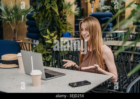 Young woman gestures and smiles while talking to laptop screen at outdoor cafe. Virtual team collaboration, executive video briefing, talking to Stock Photo