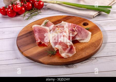 Delicate slices of cured meat, possibly prosciutto ham, arranged on rustic wooden cutting board. Fresh rosemary sprigs, vibrant red cherry tomatoes, g Stock Photo