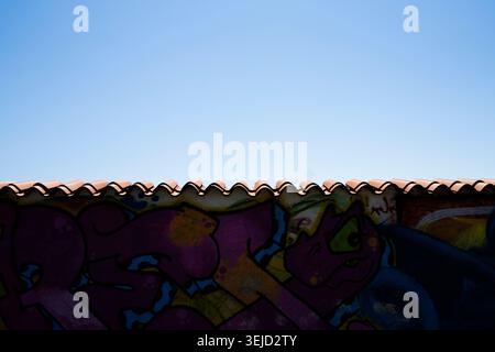 Piran, Slovenia. May 26, 2026.  A section of a tiled roof forms a wavy line against a clear blue sky. Below the roof, a wall is covered in brightly co Stock Photo