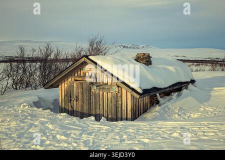Enontekiö, Finland - 05.04.2023: Rustic log cabin buried in deep snow under an overcast sky in the Lapland tundra landscape Stock Photo