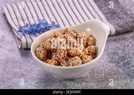 White bowl filled with delicious sesame coated nuts, likely caramelized peanuts, rests on gray textured surface. Crisp white golden brown snack presen Stock Photo