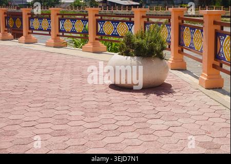 Pink hexagonal pavers line a paved walkway. Large rounded planter holds a green shrub. Colorful railing with yellow and blue motifs lines the path. Orange posts support the decorative fence Stock Photo