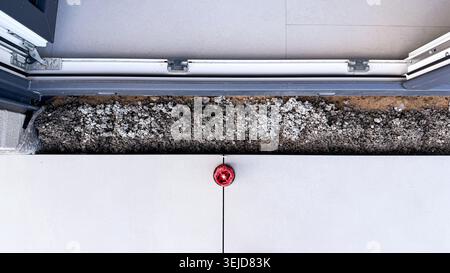 Red fire alarm bell mounted on a concrete wall near an open door with exposed gravel and flooring Stock Photo