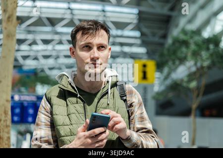 Young male passenger holding smartphone while waiting in airport terminal before flight Stock Photo