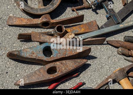 Rusted antique iron pickaxe heads and heavy hand tools are scattered on gray concrete pavement. Sunlight highlights the rough texture of the tools and Stock Photo
