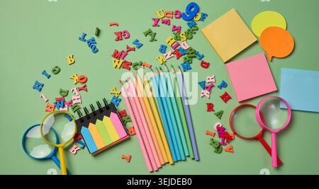 Stationery laid out on a green background. Multi-colored pencils ,  sticky notes and wooden letters, preparing for study or work Stock Photo