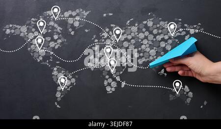 Hand holding blue paper airplane against world map background with dotted lines connecting different locations, global travel or Stock Photo