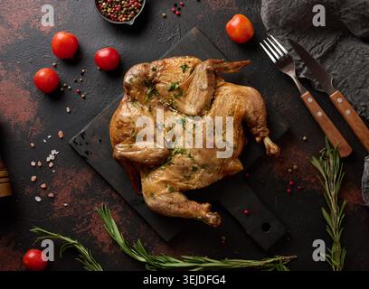 Baked chicken on a cutting board with rosemary, cherry tomatoes and spices. Dark background, wooden cutlery and kitchen utensils Stock Photo