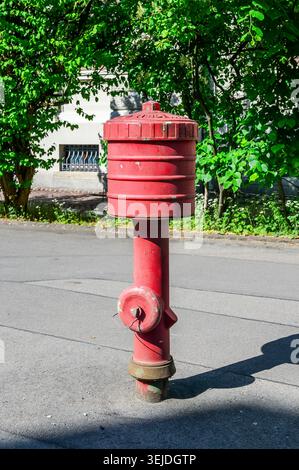 Bright red fire hydrant stands tall on a paved street with lush green trees behind Stock Photo