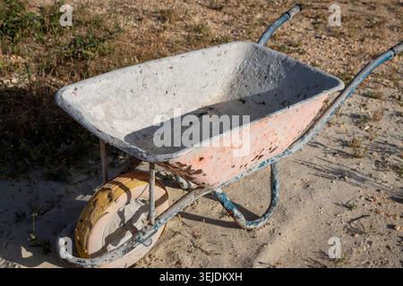 A single-wheeled cart with a worn metal basin and rusty orange exterior rests on a dry, sandy surface with sparse vegetation Stock Photo