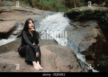 Young Asian woman sitting barefoot on rock by forest waterfall showing thoughtful peaceful expression wearing black clothing surrounded by beautiful w Stock Photo