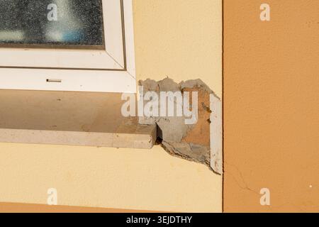 Close-up view of a building exterior revealing significant damage to the plasterwork around a window frame, exposing underlying materials and structur Stock Photo