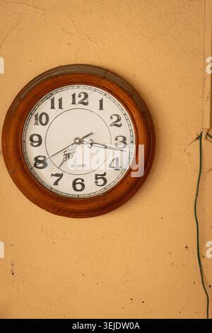 An old-fashioned round clock with a wooden frame hangs on a textured ochre wall, displaying the time with black Arabic numerals Stock Photo