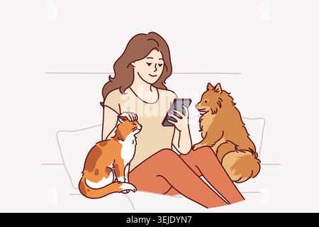 Woman owner of dog and cat sits on couch and plays on phone, enjoying spending time with pets Stock Photo